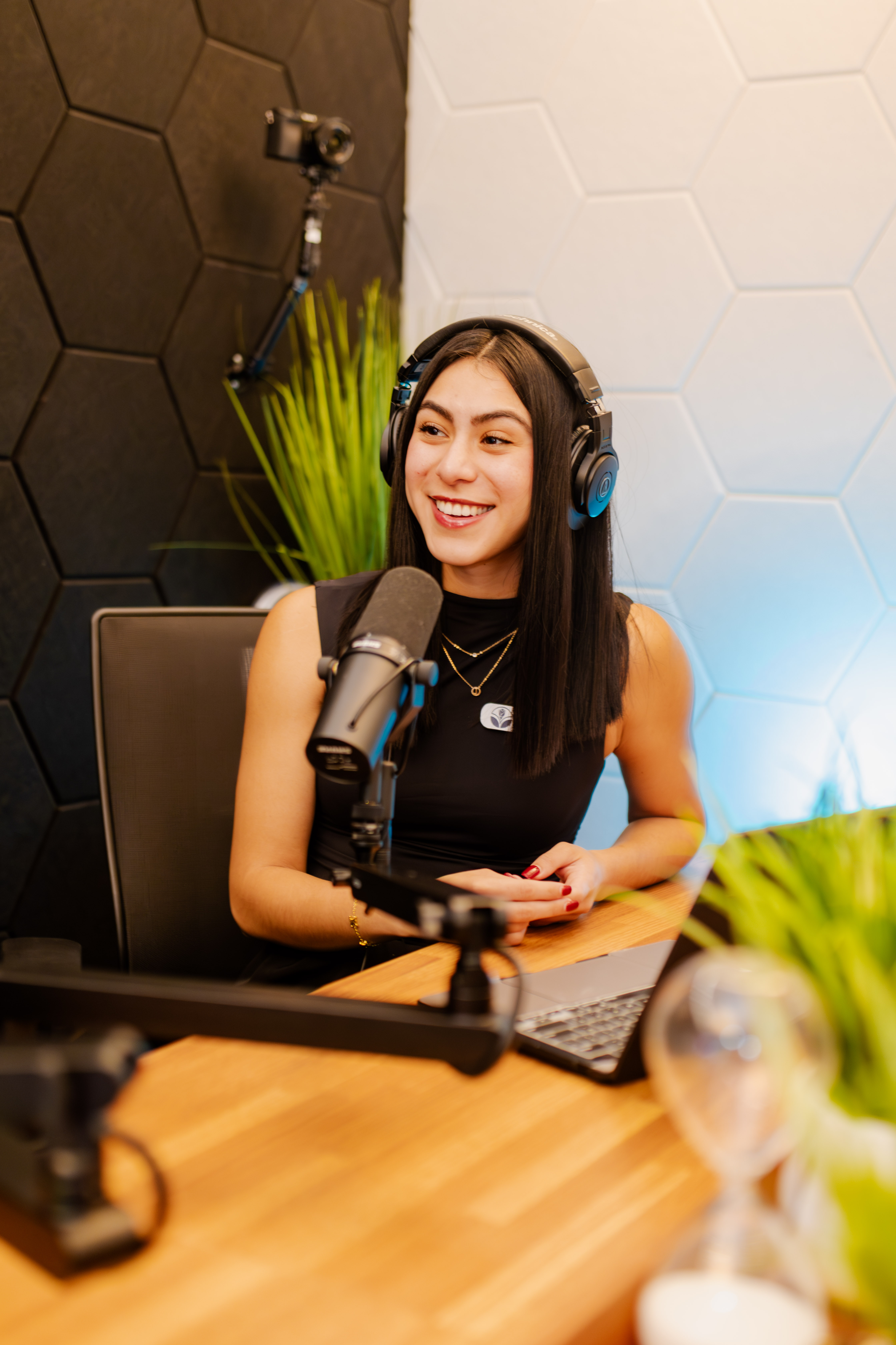 A Peachtree Rose Marketing host recording a video podcast episode in the San Antonio studio, smiling at a studio microphone with a laptop and acoustic hexagon wall panels.